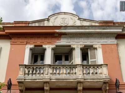 Da qui riusciamo a scorgere meglio i decori della facciata. Nella parte centrale spicca il balcone in pietra, oltre il quale intravediamo quattro bianche colonne scanalate...